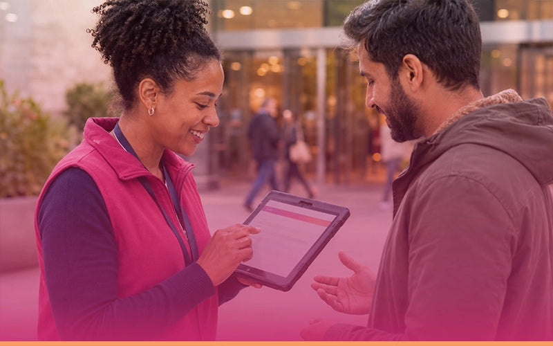 A woman in a pink vest holding a tablet and smiling at a man in a brown hoodie in an outdoor urban setting.