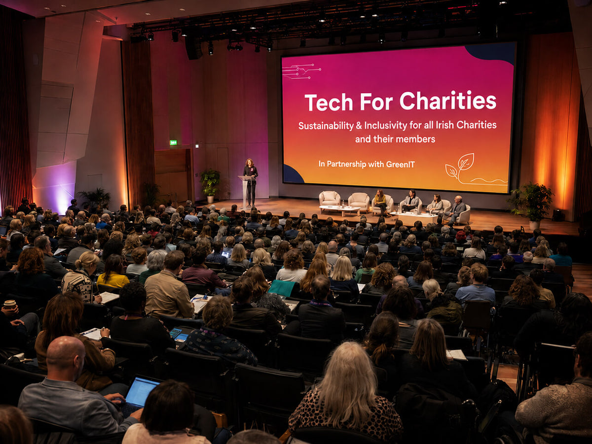 A large audience seated in rows at an auditorium, facing a stage with a speaker and a large presentation screen reading 'Tech For Charities' and 'Sustainability & Inclusivity for all Irish Charities and their members.'.