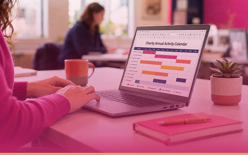 A person in a pink sweater uses a laptop on a pink table in a bright office or cafe setting. The laptop screen shows a Google Calendar-style chart titled 'Charity Annual Activity Calendar.