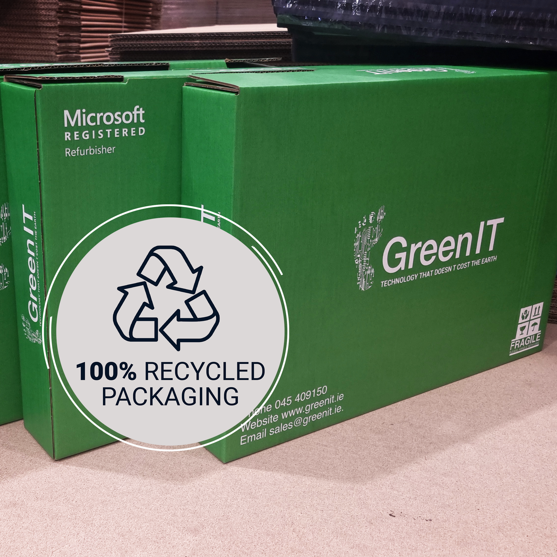Green cardboard boxes for Microsoft refurbished hardware, featuring the GreenIT logo and a large circular graphic that reads '100% Recycled Packaging' with a recycling symbol.
