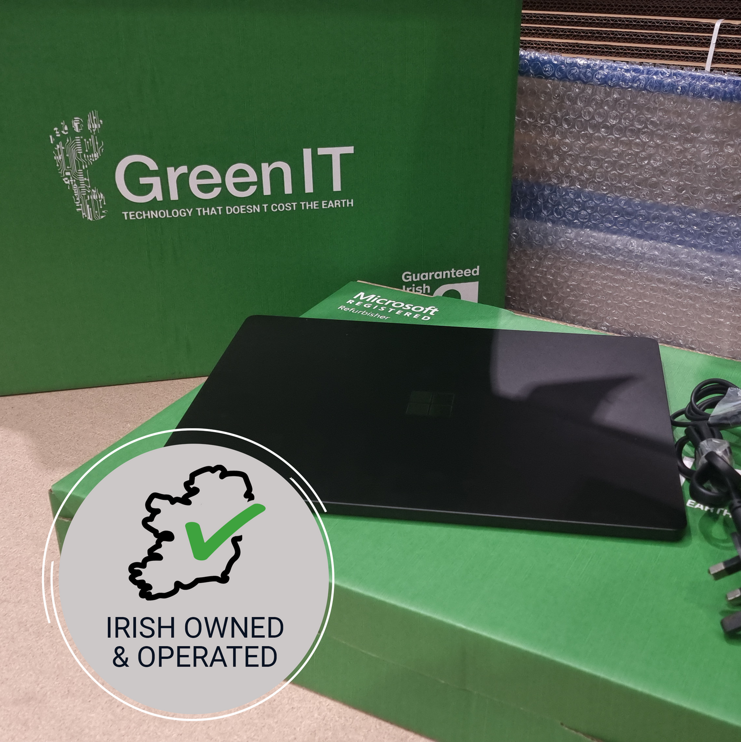 A black Microsoft Surface laptop sits on a green Microsoft Surface box, with a large green GreenIT box behind it. A circular badge in the foreground reads 'IRISH OWNED & OPERATED' with a green checkmark.