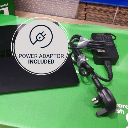 A black power adapter with a UK three-pin plug and attached power cord, placed on a bright green surface next to a black laptop. A large circular overlay reads 'POWER ADAPTOR INCLUDED' with a power icon.