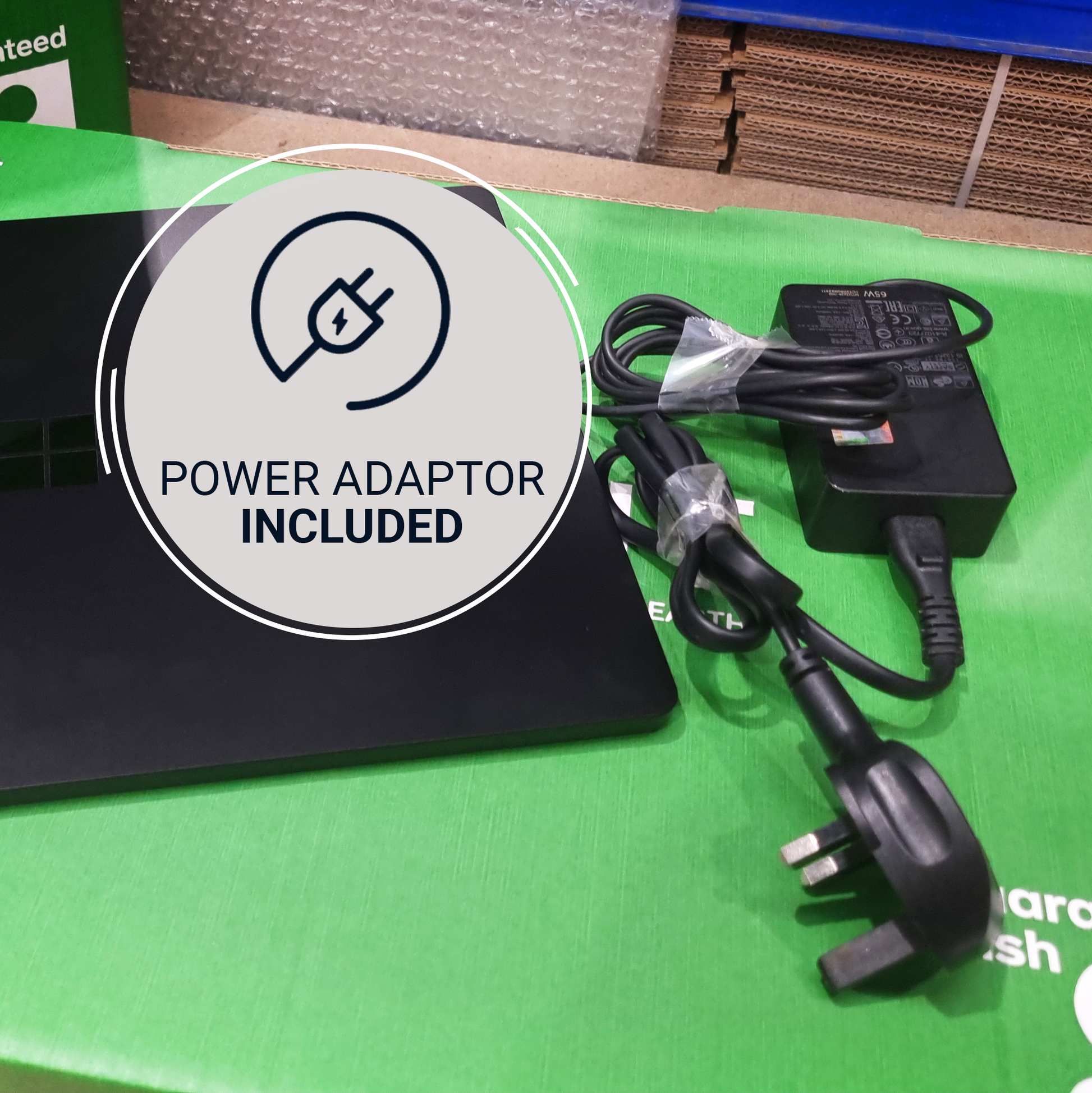 A black power adapter with a UK three-pin plug and attached power cord, placed on a bright green surface next to a black laptop. A large circular overlay reads 'POWER ADAPTOR INCLUDED' with a power icon.
